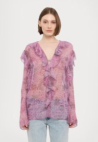 Caurspīdīga rozā blūze ar volāniem un ziedu rakstu. Ar V veida izgriezumu un garām piedurknēm, kopā ar gaiši zilām džinsa biksēm.