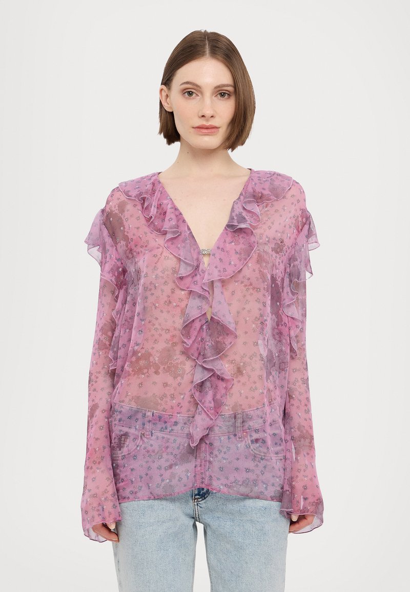 Caurspīdīga rozā blūze ar volāniem un ziedu rakstu. Ar V veida izgriezumu un garām piedurknēm, kopā ar gaiši zilām džinsa biksēm.