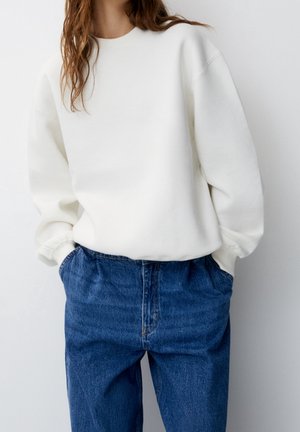 Femme portant un sweat-shirt blanc ample et un jean en denim bleu taille haute, les mains dans les poches, sur un fond uni.