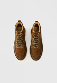 Braune Wildleder-Wanderschuhe mit goldenen Metallösen, beigen gepolsterten Kragen und cremfarbenen Gummisohlen. Verfügt über detaillierte Nähte und Zuglaschen.