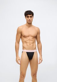 Man zonder shirt staat rechtop met het gezicht naar voren, draagt zwarte onderbroek met een bleekroze Calvin Klein tailleband tegen een effen lichte achtergrond.