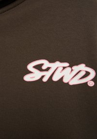 Brunt tekstil med en fremtrædende hvid og lyserød broderet logo "STWD" på fronten. Materialet virker blødt med en let struktureret overflade.