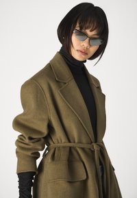 Abrigo verde oliva con cintura ceñida, cuello de solapa y grandes bolsillos; superpuesto a un cuello de tortuga negro con mangas enrolladas.