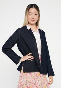 Tamno plavi traper jakna s dugim rukavima, ovratnikom i kopčanjem na gumbe. Sadrži kontrastne šavove i ležeran kroj.