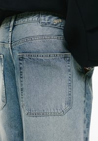 Svijetloplave traperice s visokim strukom, klasičnim džepovima i izvezenim logom blizu struka. Ujednačena tekstura i šivane šavove.