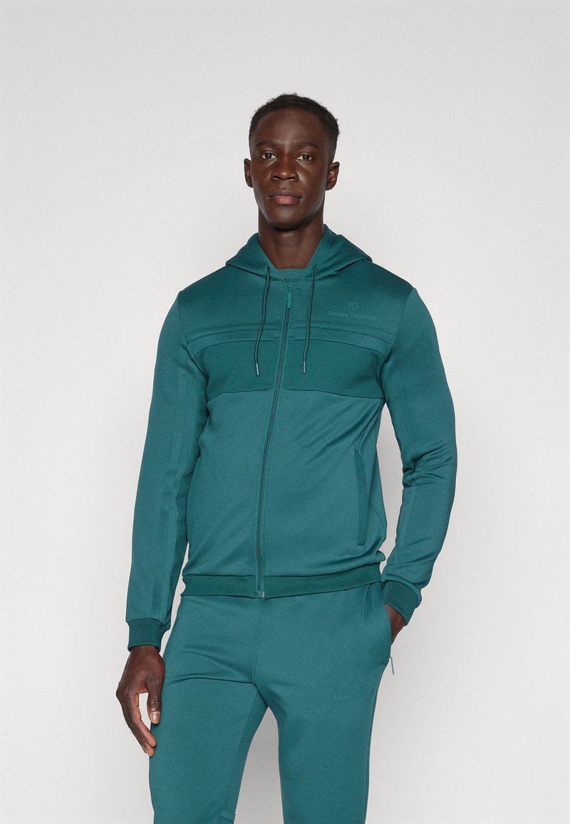 Sweat à capuche zippé teal avec un panneau texturé, capuche réglable avec cordon de serrage et poignets ajustés. Associé à un jogger teal assorti.