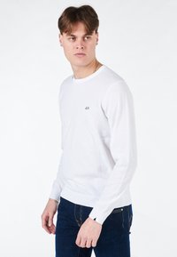 T-shirt bianca a maniche lunghe con scollo rotondo, realizzata in tessuto leggero, con un piccolo logo nero sul petto, abbinata a jeans scuri.