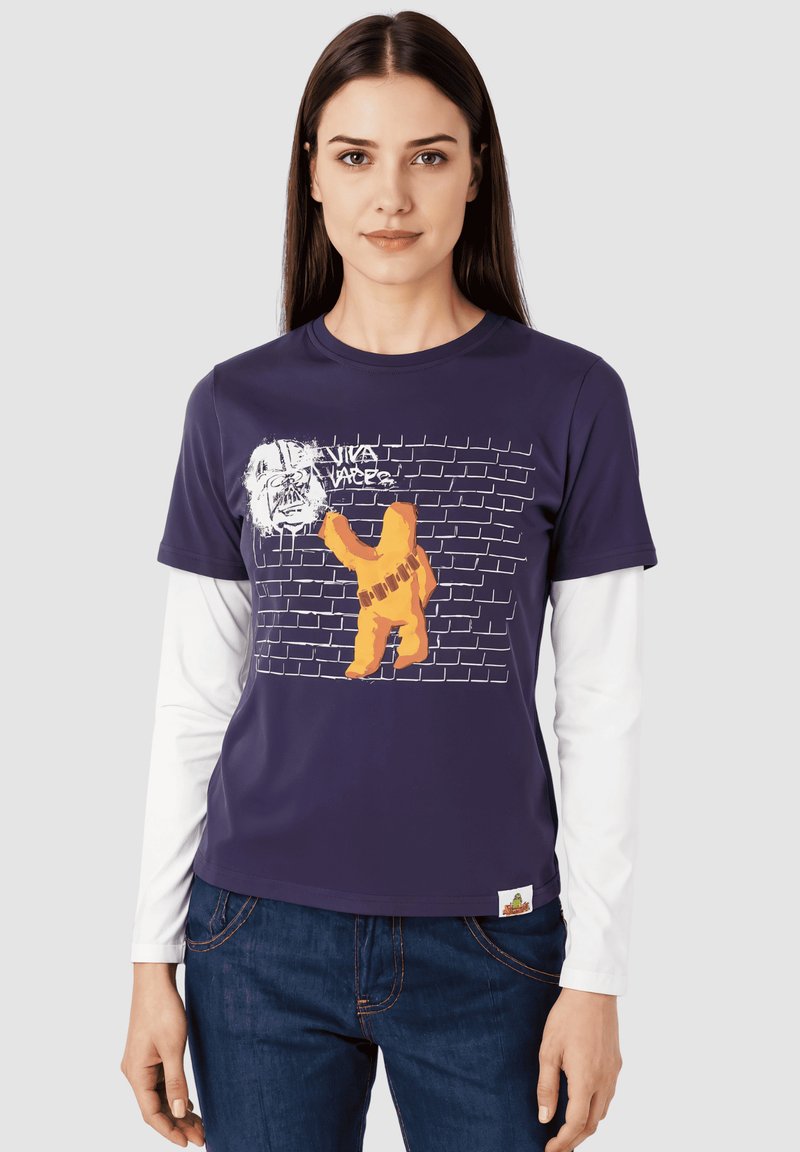 Camiseta marina con un gráfico de un personaje naranja sobre una pared de ladrillos, con texto blanco y mangas largas blancas debajo. Material de mezcla de algodón.