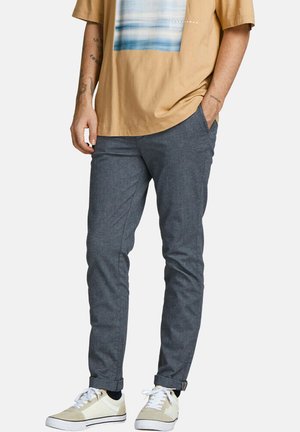 Pantalons gris cintrés en tissu texturé, avec des poches latérales et un motif subtil, associés à des baskets blanches et un t-shirt graphique beige.