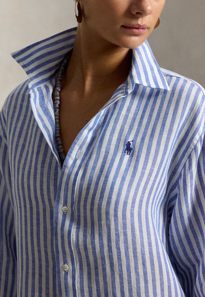 Kvinne iført blå og hvit vertikalt stripete skjorte med knapper, krage stikkes ut og perlekjede, kuttet ved hake og skulder.