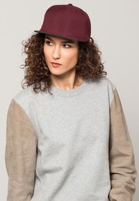 Gorra burdeos con visera plana, material texturizado y un color verde contrastante en el interior. Llevada con un sudadera gris que presenta mangas de ante color beige.