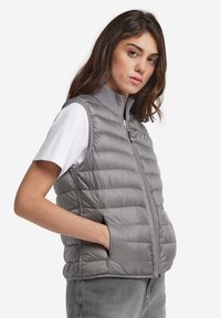 Jasnoszara kamizelka puchowa z wysokim kołnierzem, z poziomym przeszyciem, zamek błyskawiczny z przodu oraz bocznymi kieszeniami. Nosi się ją na białej koszuli.