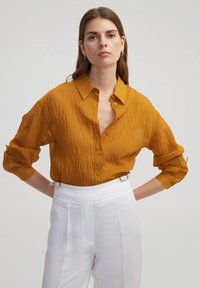 Senfgelbes Button-Down-Hemd aus gekrauster Stoffqualität, mit Kragen, hochgerollten Ärmeln und einer Brusttasche. Getragen mit weißen, hoch taillierten Hosen.