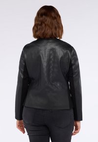 Donna con i capelli corti castani, che indossa una giacca nera trapuntata e jeans scuri, in piedi di spalle contro uno sfondo chiaro uniforme.