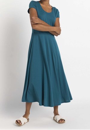 Woman standing with arms crossed wearing a teal short-sleeve midi dress and white slide sandals against a plain background.