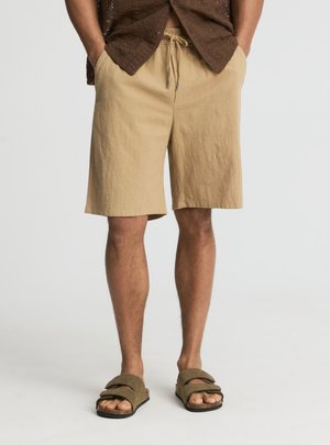 Mężczyzna w beżowych szortach ze sznurkiem, brązowych sandałach i brązowej teksturowanej koszuli zapinanej na guziki, z rękami w kieszeniach.
