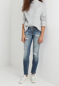 Pull encolure ras du cou gris à côtes associé à un jean bleu clair déchiré et des baskets blanches. Présente une coupe décontractée et des déchirures visibles dans le jean.