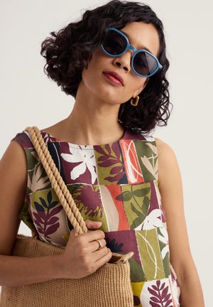 Moteris, dėvinti mėlynus akinius nuo saulės ir be rankovių gėlėtą siuvinėtą suknelę, laikanti rudos spalvos juostinę rankinę su storomis virvės rankenomis.