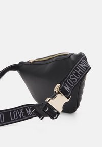 Černá texturovaná taška se zlatým zipem, vybavená výrazným nastavitelným popruhem s bílým textem a kovovou sponou. Trojúhelníkový tvar.