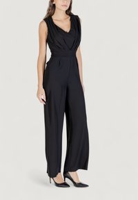 Mono negro sin mangas con escote drapeado en forma de V, pierna ancha y cinturilla ajustada. Tela suave con un sutil brillo, combinado con tacones de punta.