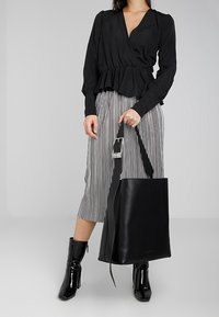 Kobieta w czarnej bluzce i spódnicy w paski trzyma dużą czarną skórzaną torbę na ramię, do tego błyszczące czarne botki.