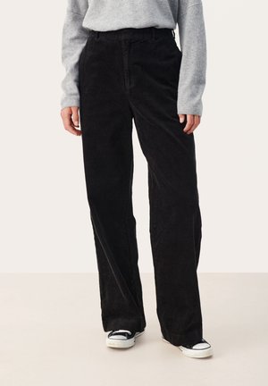 Personne portant un pantalon large en velours côtelé noir, un pull gris à manches longues et des baskets noir et blanc, debout sur un fond uni.