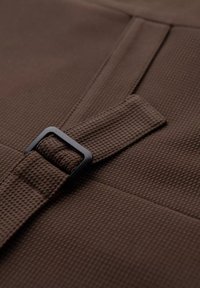 Braunes, strukturiertes Material mit einem gewebten Muster, das eine schwarze rechteckige Schnalle zeigt, die einen Riemen sichert. Das Material wirkt robust und strapazierfähig.