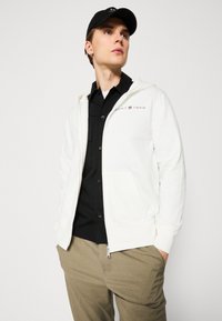 Felpa bianca con zip e tasche, con un logo rosso e grigio che recita "GANT 1949." Indossata sopra una camicia nera e pantaloni beige.