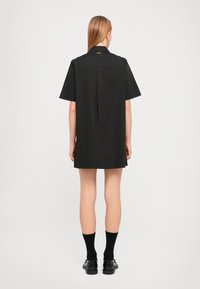 Black short-sleeve dress with a straight cut, subtle back pleat, and a gold logo detail on the collar. Worn with black socks and shoes.