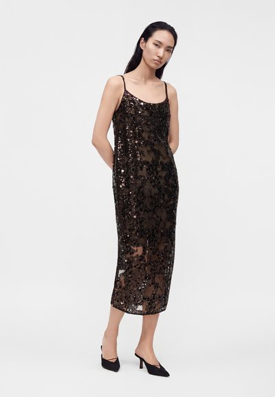 Femme portant une robe midi noire sans manches à sequins avec des fines bretelles et des escarpins noirs à bout pointu, posant devant un fond uni.