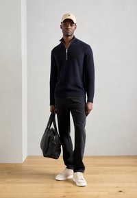 Marineblå lynlås sweater, sorte jeans og hvide sneakers. En sort taske holdes af personen, som bærer en beige kasket med et logo.