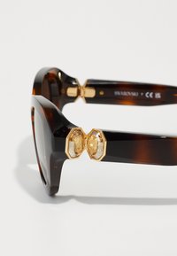 Schildpatt-Sonnenbrille mit auffälligen hexagonalen goldenen Akzenten und klaren Steinen an den Scharnieren. Glänzende Oberfläche, markantes Rahmendesign.