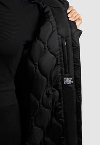 Sort pufferjakke med quiltet tekstur og indvendig lomme. Har brandlogo på indersiden. Nærbillede, der viser ærmet og hånd, der holder stoffet.