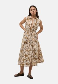 Robe sans manches à motif floral marron et blanc, avec un col et une taille à cordon. Longueur en dessous du genou, accompagnée de sandales sombres.