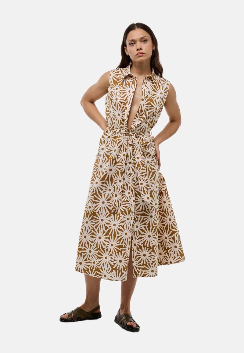 Robe sans manches à motif floral marron et blanc, avec un col et une taille à cordon. Longueur en dessous du genou, accompagnée de sandales sombres.