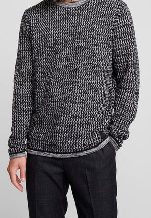 Hombre con un suéter de punto texturizado en blanco y negro y pantalones oscuros a medida, con una mano en el bolsillo, frente a un fondo liso.