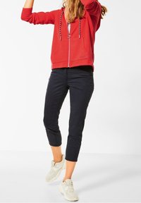 Felpa rossa con zip e coulisse, abbinata a pantaloni neri cropped e sneakers chiare, che mostrano uno stile sportivo casual.