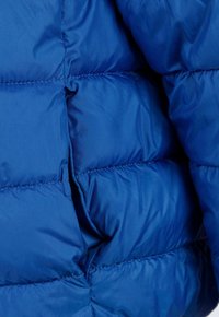 Blå puffjacka med quiltade sektioner och glänsande syntetiskt tyg, med en mjuk yta och synliga sömnadsdetaljer genom hela plagg.