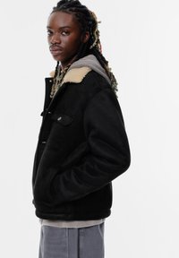 Czarna, strukturalna kurtka z beżowym futrzanym kołnierzem i przednimi zatrzaskami, nałożona na szary hoodie. Model pozuje w profilu na tle białego tła.
