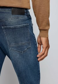 Tmavě modré džíny z denimu s vybledlou texturou, s jednou zadní kapsou a černou koženou značkovou nášivkou u pasu.