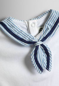 Bluza albă are un guler de marinar cu dungi albastre și albe, având un accent de albastru închis, fixat cu un nasture la spate.