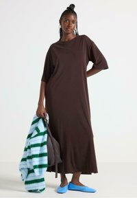 Brun, knælang kjole med korte ærmer, lavet af et blødt stof. Komplementeret af en stribet blå og grøn sweater og blå flade sko.