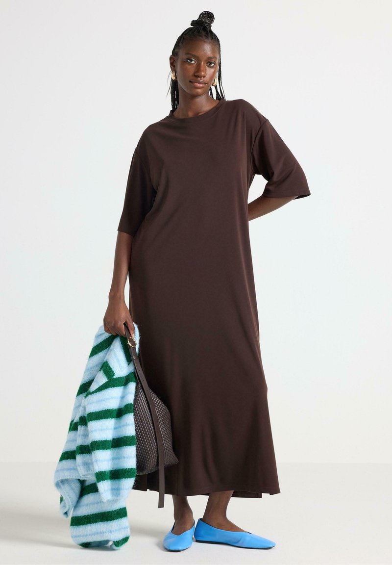 Brun, knælang kjole med korte ærmer, lavet af et blødt stof. Komplementeret af en stribet blå og grøn sweater og blå flade sko.