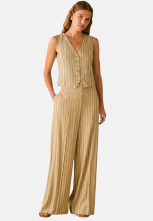 Donna che indossa un gilet beige a righe sottili senza maniche con bottoni e pantaloni a gamba larga con le mani nelle tasche, su sfondo semplice.