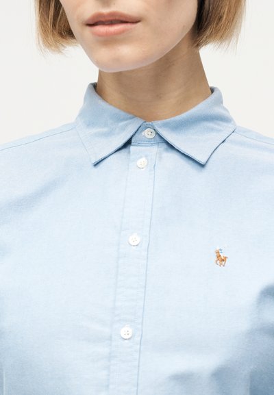 Γυναίκα που φοράει ένα ανοιχτό μπλε πουκάμισο με κουμπιά και μικρό κεντημένο λογότυπο παίκτη πόλο στο αριστερό στήθος.