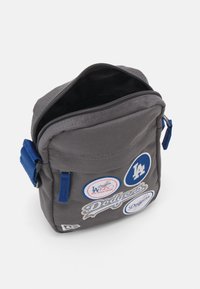 Borsa a spalla in tessuto grigio con accenti blu, dotata di numerosi patch dei Dodgers e del logo di Los Angeles. Presenta una chiusura con zip e una tracolla regolabile.
