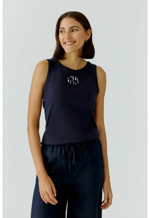 Femme souriante aux cheveux bruns mi-longs portant un haut noir sans manches et un pantalon noir à cordon de serrage, sur un fond uni.