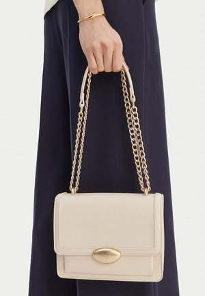 Hand, die eine beige rechteckige Handtasche mit goldener Kettenriemen und ovalem goldfarbenem Verschluss hält, vor dunklem Marinehosen-Hintergrund.