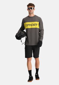 Grijze langemouwshirt met een gele horizontale streep en "SPYDER" logo, zwarte shorts, zwarte handschoenen en een helm in de hand. Zwarte schoenen en sokken.