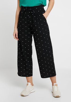 Kobieta ubrana w czarne szerokie, krótkie spodnie w białe kropki, zielony top i białe trampki, ręce w kieszeniach.